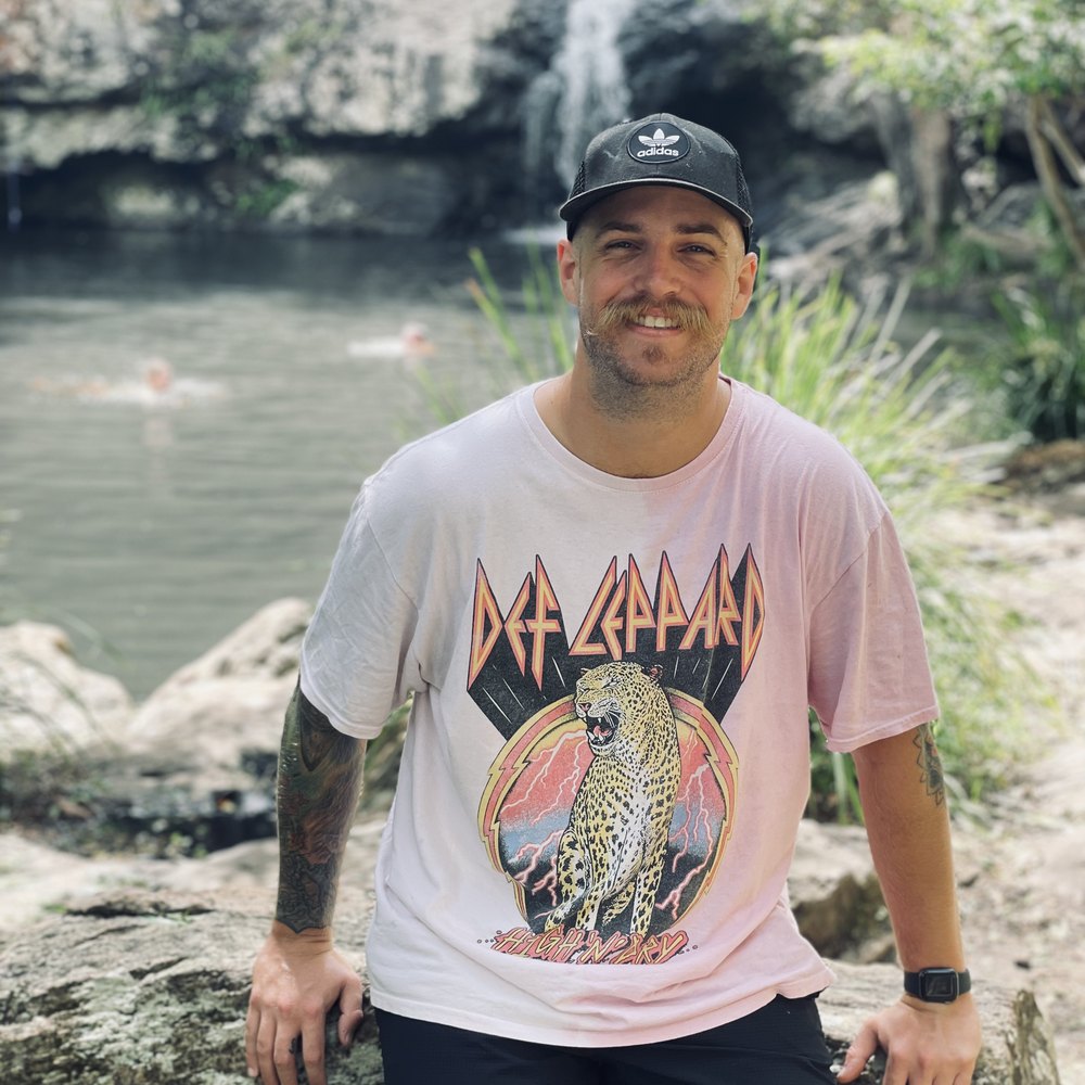 Luke smiling in a black cap and pink tee, sitting on a rock by a waterfall